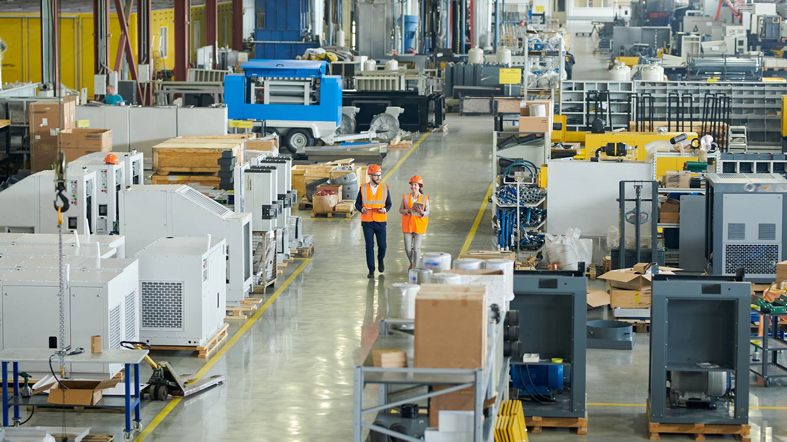 High angle full length portrait of bearded businessman wearing hardhat walking across production workshop accompanied by female factory employee