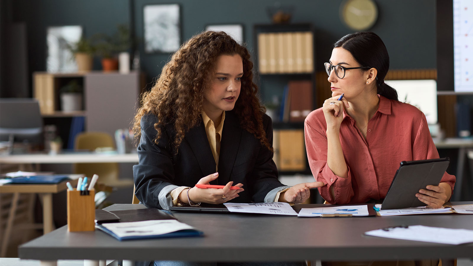 Two young adult women sitting at a desk discussing documents and digital tablet in modern office workspace